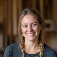 Portræt af Sofie Andersen, en smilende møbelsnedker i sit værksted, omgivet af træspåner og værktøj, i naturligt lys.